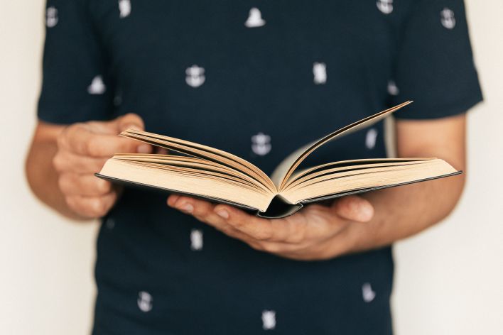 Anonymous male in dark tee shirt reading interesting book while standing in front of light wall